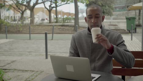 Young-man-in-coat-using-laptop-and-drinking-from-paper-cup